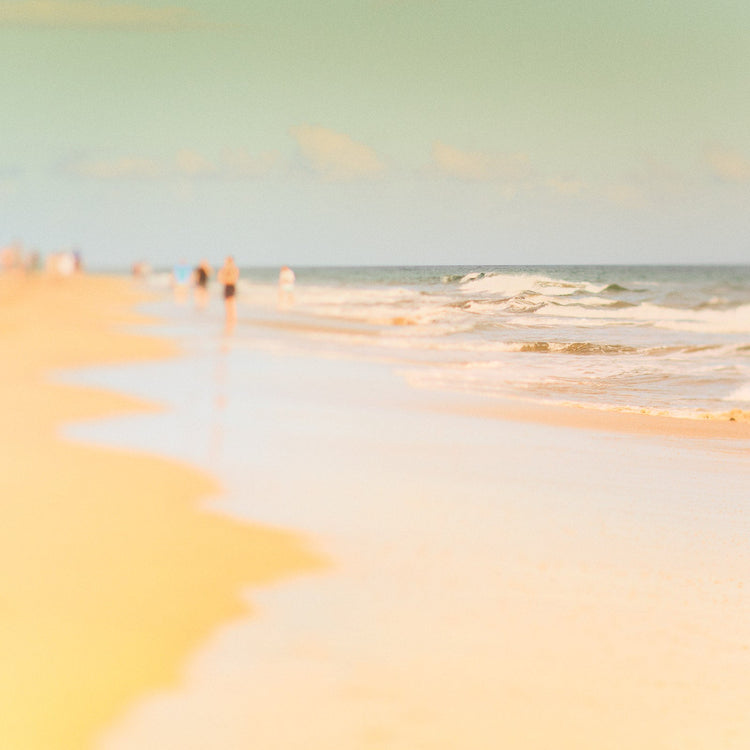 Ein quadratisches Bild von Wellen, die an einen Strand fliessen und unscharf Menschen im Hintergrund
